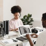 Dois profissionais colaboram em um escritório moderno, usando laptops ao lado de equipamentos de câmera.
