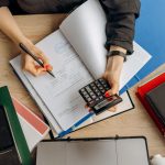 Profissional analisa documentos financeiros com uma calculadora sobre a mesa, em um contexto de trabalho contábil.