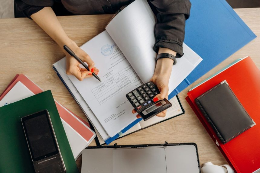 Profissional analisa documentos financeiros com uma calculadora sobre a mesa, em um contexto de trabalho contábil.