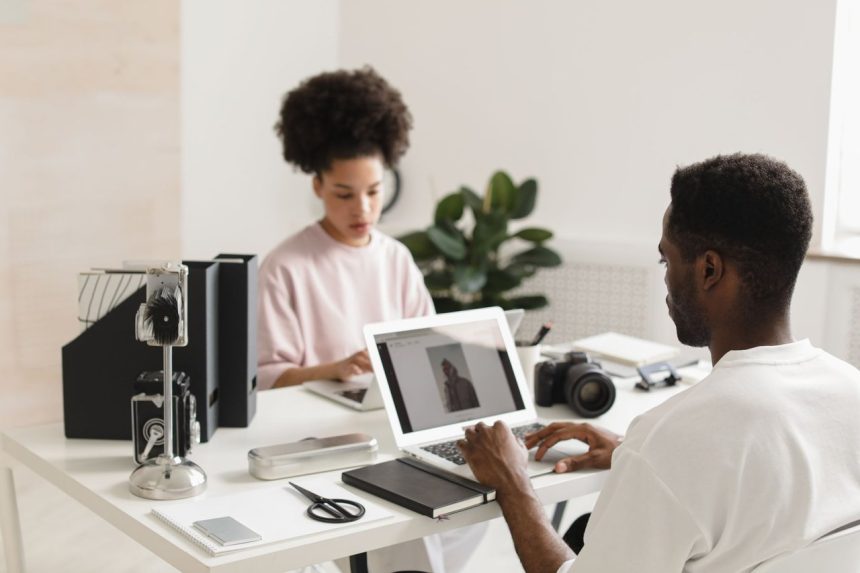 Dois profissionais colaboram em um escritório moderno, usando laptops ao lado de equipamentos de câmera.