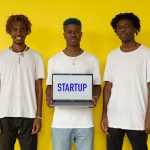 Três homens seguram um notebook com a palavra “STARTUP” diante de um fundo amarelo.