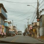Rua comercial movimentada, com lojas, veículos e pedestres em um dia nublado.