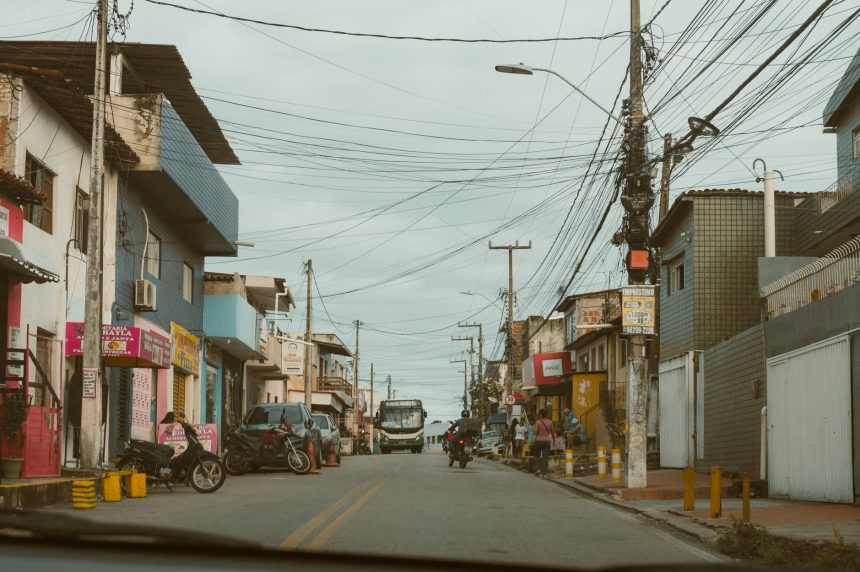 Rua comercial movimentada, com lojas, veículos e pedestres em um dia nublado.