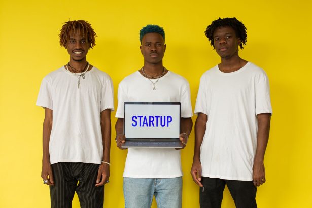 Três homens seguram um notebook com a palavra “STARTUP” diante de um fundo amarelo.