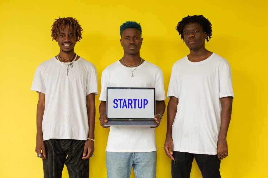Três homens seguram um notebook com a palavra “STARTUP” diante de um fundo amarelo.
